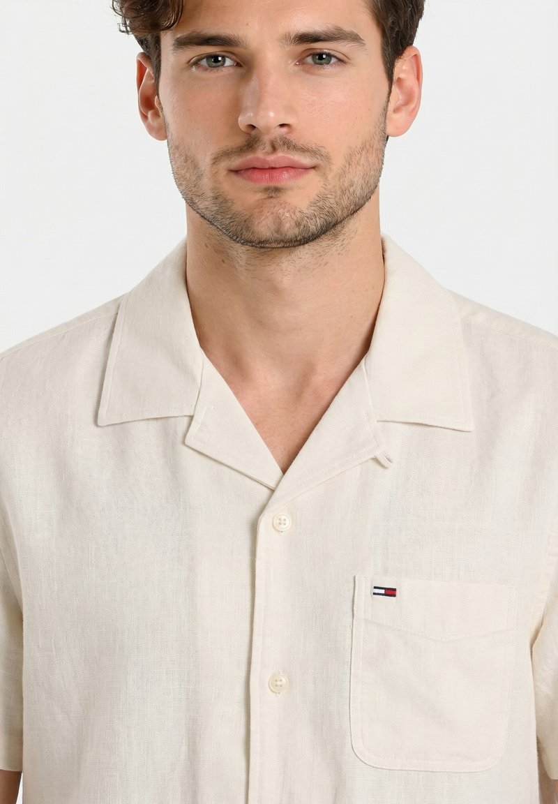 Jeune homme aux cheveux courts et bruns portant une chemise crème à manches courtes avec une petite poche poitrine et un logo.