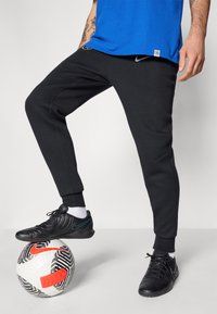 Sorte sweats med tapered ben, lavet af bomuldsstof, og elastiske manchetter. Båret med sorte fodboldsko, stående på en mønstret fodbold.