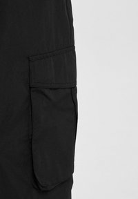 Schwarze Cargo-Shorts aus Stoff verfügen über eine große Seitentasche mit Klappe, genähten Kanten und einer glatten Textur. Minimalistisches Design mit funktionalen Details.