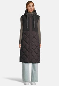 Femme portant un long gilet matelassé sans manches noir avec une capuche sur un pull col roulé gris, un jean large bleu clair et des baskets blanches.