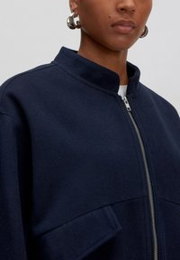 Marineblaues Zip-Jackett mit Stehkragen, aus strukturiertem Stoff gefertigt. Verfügt über eine Seitentasche und einen silbernen Reißverschlussakzent.