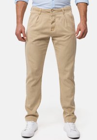 Chinos beiges avec plis à l'avant, fermeture éclair et bouton. Comprend des poches latérales et des jambes droites, associés à des baskets blanches.