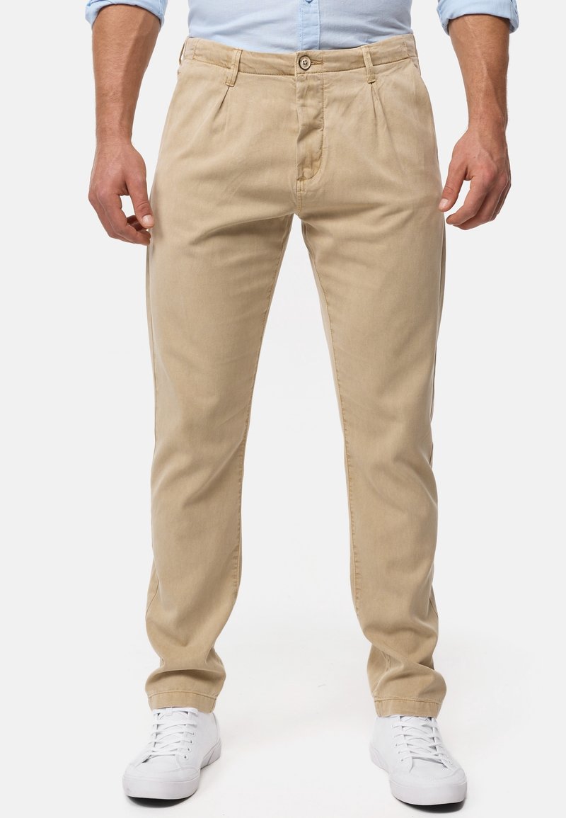 Chinos beiges avec plis à l'avant, fermeture éclair et bouton. Comprend des poches latérales et des jambes droites, associés à des baskets blanches.