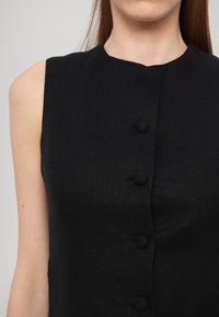Černé šaty bez rukávů s kulatým výstřihem a pěti knoflíky potaženými látkou vpředu, vyrobené z texturovaného materiálu.