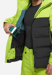 Eine grüne und schwarze isolierte Jacke mit einer Reißverschlusstasche. Eine Person hält Ski-Goggles und trägt eine schwarze Basisschicht sowie passende Hose.