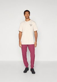 Smėlio spalvos medvilniniai marškinėliai su nedideliu purpuriniu grafiniu simboliu ir bordo spalvos laisvalaikio kelnės. Kelnėse yra gofruoti manžetai ir šoninės kišenės. Juodi sportiniai bateliai.