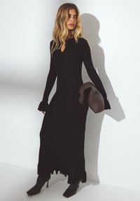 Robe noire longue à côtes avec des manches longues, décolleté en trou de serrure et ourlet volanté ; associée à une pochette tressée marron et des escarpins noirs à bout pointu.