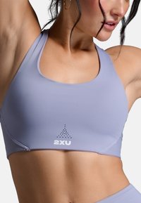 Svetlofialová športová podprsenka vyrobená z hladkého, elastického materiálu. Má logo na prednej strane a dizajn s prekríženými ramienkami pre pohodlný pohyb.