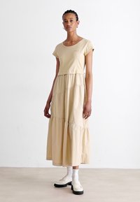 Beige katoenen maxi-jurk met korte mouwen, tiered ruffle ontwerp en een relaxed fit. Gecombineerd met witte sokken en chunky schoenen.