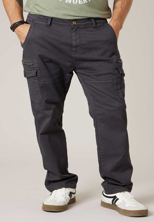 Mann trägt dunkelgraue Cargo-Hose mit Seitentaschen und weiße Turnschuhe mit schwarzen Details, steht mit Händen in den Taschen.