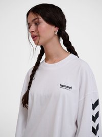 Fiatal nő fonott hajjal, fehér Hummel sportmelltartót visel, fekete cikk-cakk csíkokkal az ujjon, balra néz.