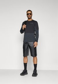 Haut à manches longues noir avec des accents gris, shorts noirs brillants, chaussettes noires et chaussures ; modèle portant un sac à dos et des lunettes de soleil.