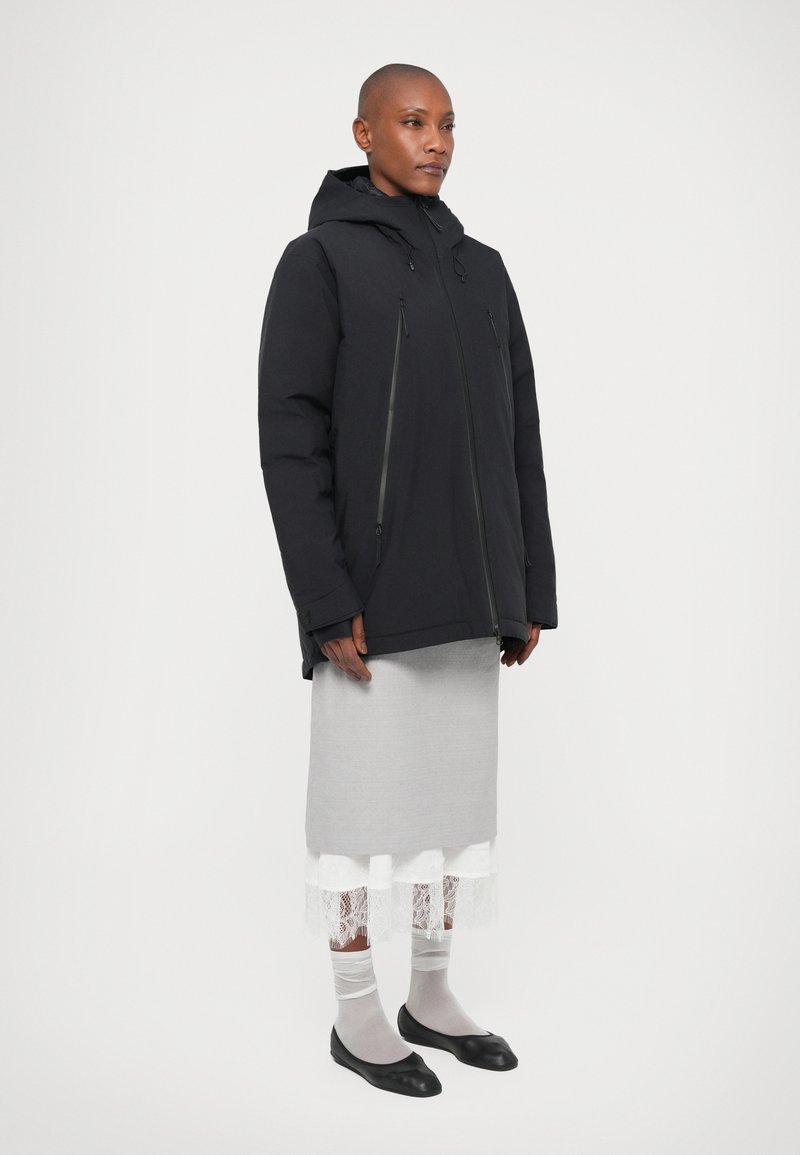Veste noire matelassée avec poches zippées et capuche, associée à une jupe grise arrivant aux genoux avec une bordure en dentelle blanche et des ballerines noires.