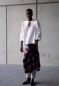 Blusa branca de mangas bufantes com um decote redondo e detalhe em buraco de chave, combinada com calças cinzas estampadas e um xale xadrez em vermelho e azul. Sapatos pretos.