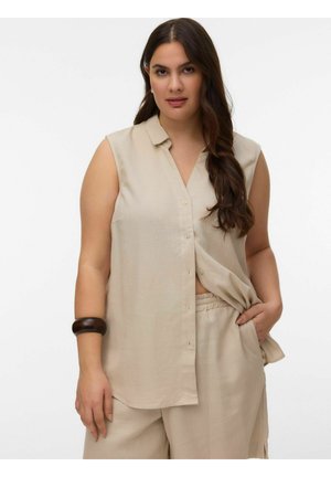 Kvinde med langt mørkt hår iført ærmeløs beige skjorte med knapper og matchende bukser, hånd i lommen, og et mørkt armbånd på venstre håndled.