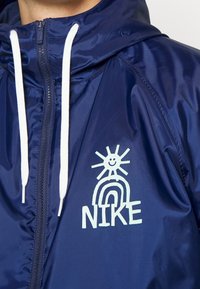 Marinblå vindjacka med vita dragsnören och en dragkedja framtill. Har en grafisk design med en leende sol och regnbåge samt texten "NIKE" i fet stil.
