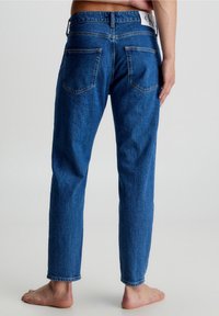 Siniset farkut suoran lahkeen muotoisina, näkyvät saumat, kaksi takataskua ja vyötärön etiketti, esitettynä takaviistosta.