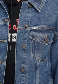 Denim jakna v srednji modri barvi z zmanjšenim učinkom, z črnimi pritisnimi gumbi in žepom na prsih. Pod njo je črna kapuca z rdečimi detajli.