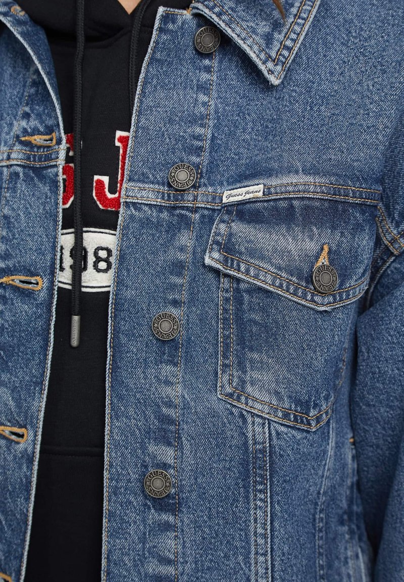 Chaqueta de denim en azul medio con textura desgastada, presenta botones a presión negros y un bolsillo en el pecho. Debajo lleva una sudadera negra con detalles en rojo.