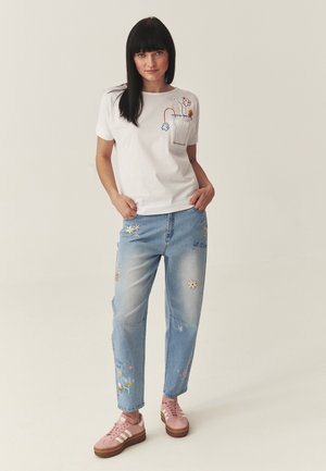 Mustajuukseline naine, kes kannab valget t-särki lillemustriga tikandiga, helerohelisi teksapükse lillekujudega ja roosasid tossusid.