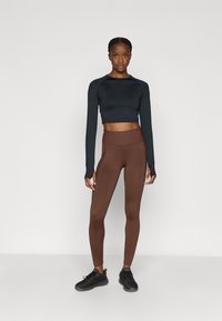 Juodas trumpintas ilgomis rankovėmis marškinėlis, derinamas su rudos spalvos aukštu liemeniu tamprėmis ir juodais sportiniais batais, pasižymintis aptemptu dizainu ir lygiu audiniu.