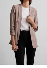 Svetle béžový blazer vyrobený z hladkého materiálu, s úzkým střihem, otevřeným předním dílem a ohrnutými rukávy, doplněný bílým topem a černými kalhotami.