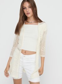 Young woman wearing white lace cardigan, white tube top, white shorts, and silver necklace, standing against plain light background.