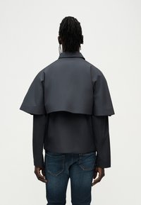 Σκούρο γκρι μπουφάν με μοναδικό σχέδιο που μοιάζει με κάπα στο πίσω μέρος, με απαλή υφή και μακριά μανίκια, σε συνδυασμό με μπλε τζιν.