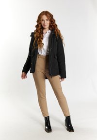 Chaqueta de plumas negra con capucha, pantalones beige ajustados y botas de tobillo negras brillantes. La chaqueta cuenta con cierre de cremallera y botones a presión.