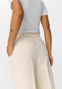 Pantalons beige à taille élastique avec poches arrière, texture lisse, design à jambe large, associés à un haut ajusté à manches courtes de couleur gris clair.