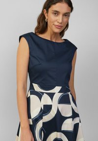 Navyblaues Kleid mit tailliertem Oberteil und kurzen Ärmeln, mit einem plissierten Rock, der mit kreisförmigen Mustern in Creme und Hellblau verziert ist.