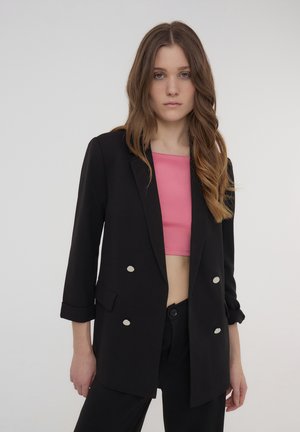 Blazer nero con bottoni argentati, abbinato a un top cropped rosa. Presenta spalle strutturate e maniche arrotolate. Tessuto morbido.