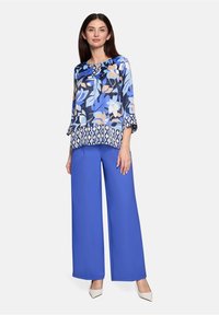 Haut bleu à motif floral avec manches 3/4, motif en couches. Associé à un pantalon bleu à jambes larges. Le mannequin pose sur un fond uni.