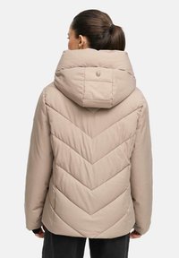 Veste matelassée beige avec capuche large et motif en chevrons au dos. Caractérisée par un petit logo sur la capuche et des poignets foncés.