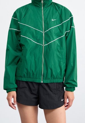 Veste zippée verte Nike avec passepoil blanc et logo, associée à un short noir Nike avec un logo blanc sur la jambe gauche.