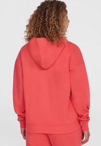 Korallenfarbener, übergroßer Hoodie mit einer verstellbaren Kapuze, gerippten Bündchen und Saum. Weicher Baumwollstoff mit glatter Textur.