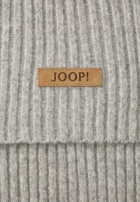 Tejido de punto gris con textura de estrías verticales, que presenta un parche de cuero marrón rectangular etiquetado "JOOP!" con letras en relieve.