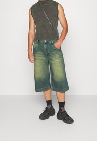Top sin mangas de canalé verde, pantalones cortos tipo Bermuda de mezclilla con degradado de claro a oscuro, y zapatillas deportivas negras con suelas texturizadas y calcetines.