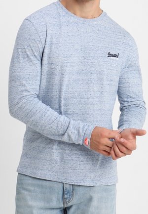 Homme portant une chemise bleu clair à manches longues avec un petit logo noir sur la poitrine et une étiquette rouge sur le poignet, ajustant la manche, associé à un jean bleu clair.