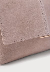 Γωνία τσάντας από καφέ σουέντ με ορατές ραφές και μικρή χρυσή μεταλλική λεπτομέρεια σε ανοιχτό φόντο.