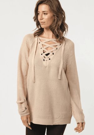 Mujer vestida con un suéter de punto beige, con escote de encaje entrecruzado y detalles de tiras a juego en los puños, mirando hacia su izquierda.