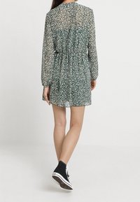 Vihreä kukallinen mekko, jossa on pitkät, läpikuultavat hihat, vyötärö kavennettu ja laskeutuva helma. Yhdistetty mustien korkeavartisten tennarien kanssa.