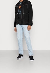 Svart denimjacka med vit sömnad över en svart grafisk t-shirt, kombinerad med ljusblå jeans och svarta sneakers med vita sulor.