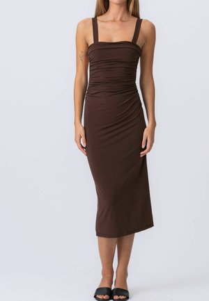 Kobieta ubrana w dopasowaną, bezrękawną, brązową sukienkę midi z szerokimi ramiączkami i marszczeniami, do której dobrała czarne klapki z odkrytymi palcami.