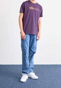 Fialové bavlněné tričko s logem "ALPHA", kombinované se světle modrými džíny a bílými teniskami, stojící na modrém kobercovém povrchu.