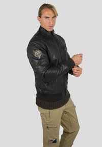 Veste en cuir noir avec poignets et taille côtelés, présentant un écusson rond sur le bras gauche, sur un fond uni.