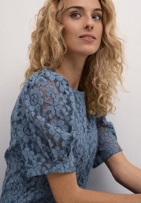 Blue lace blouse featuring floral patterns, short sleeves, and a round neckline, made of sheer, textured fabric with a relaxed fit.
