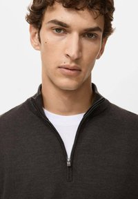 Dunkelgrauer Reißverschluss-Pullover mit hohem Kragen, feiner Strickstruktur und silbernem Reißverschluss. Wird über einem weißen T-Shirt getragen. Glattes Design und figurbetonte Form.