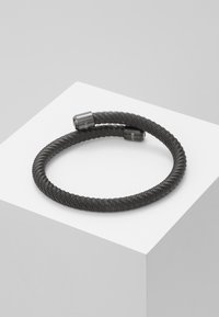 Bracciale nero avvolgente realizzato in materiale flessibile. Presenta due estremità in metallo con testo inciso, creando un design minimalista.