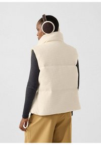 Gilet color crema, dalla texture lavorata, con collo alto, abbinato a un maglione scuro e pantaloni beige. Include cuffie imbottite marroni.